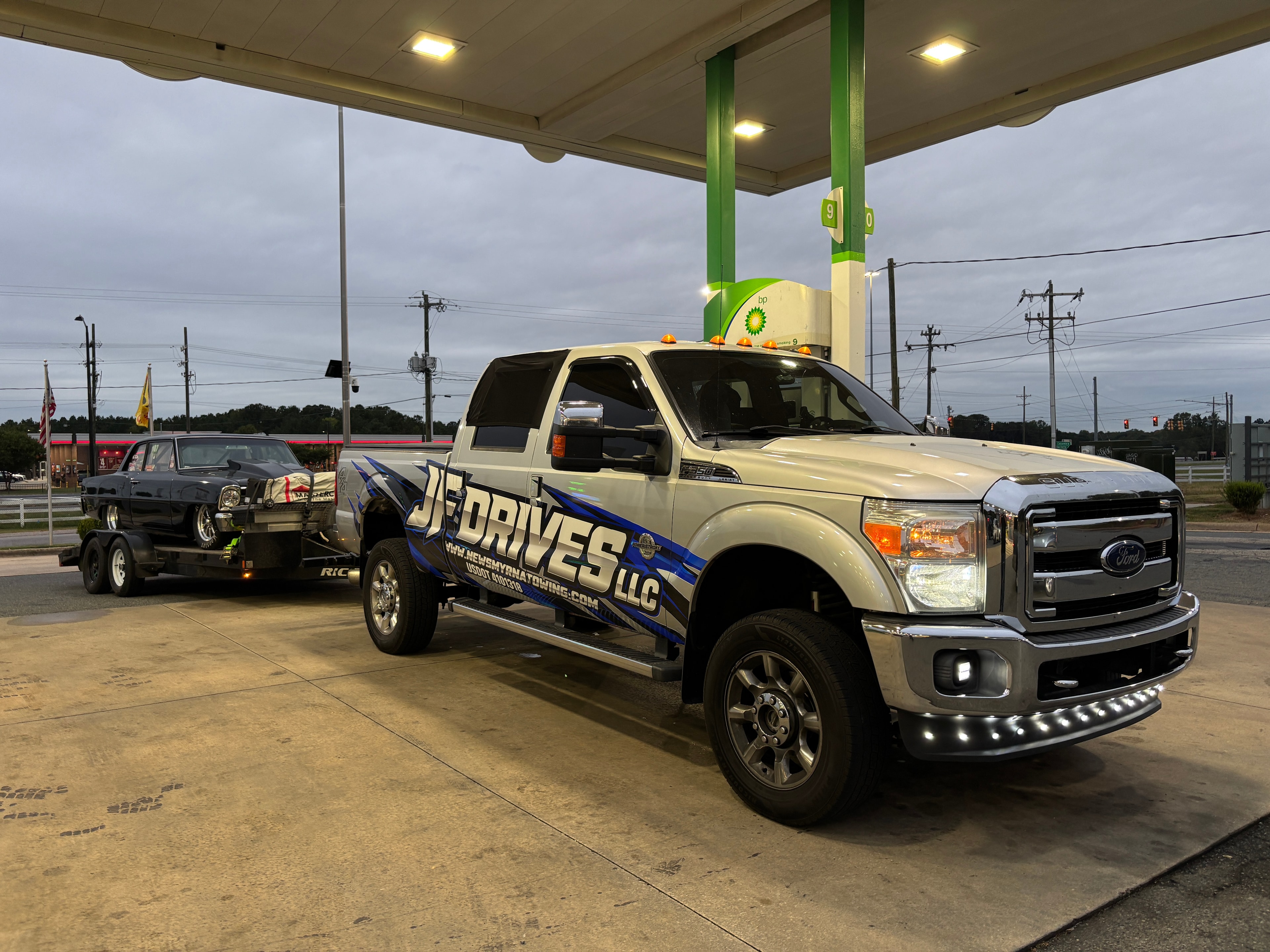 JF Drives LLC tow truck transporting a classic car at a gas station