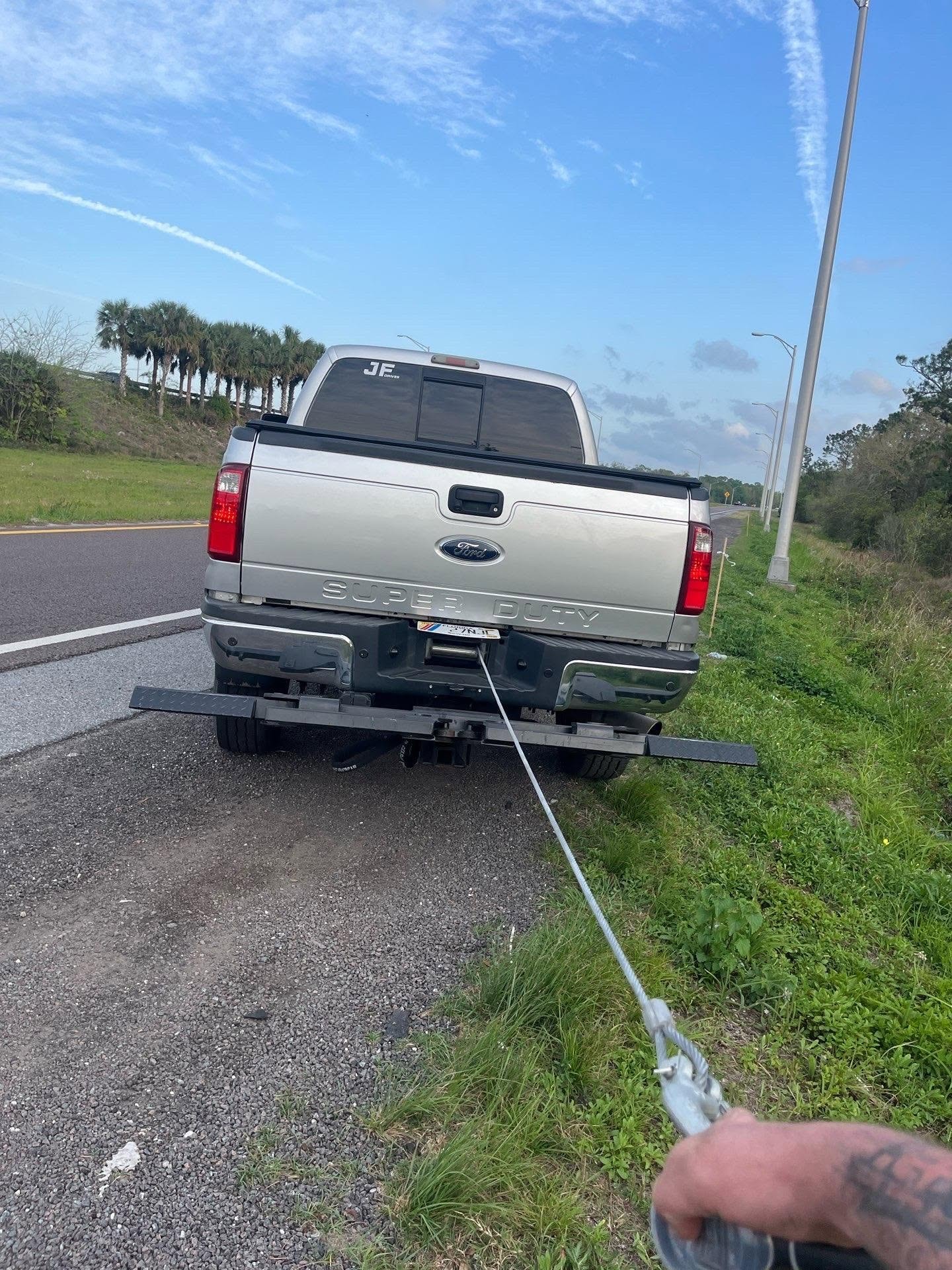 Close-up of JF Drives winch cable running from the recovery truck toward the camera during an active recovery — truck rear, cable, and hand holding the hook visible